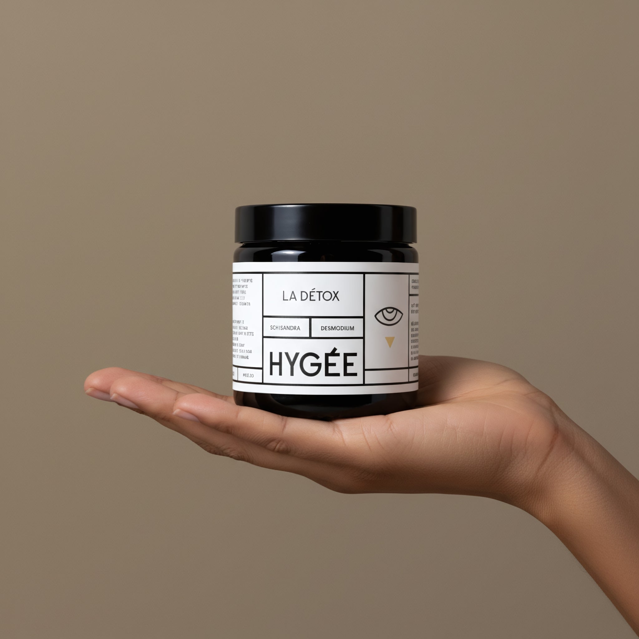 A neutral‑toned hand presents a black‑lidded amber HYGÉE La Détox jar against a smooth taupe background, showing the compact format and minimalist white label.