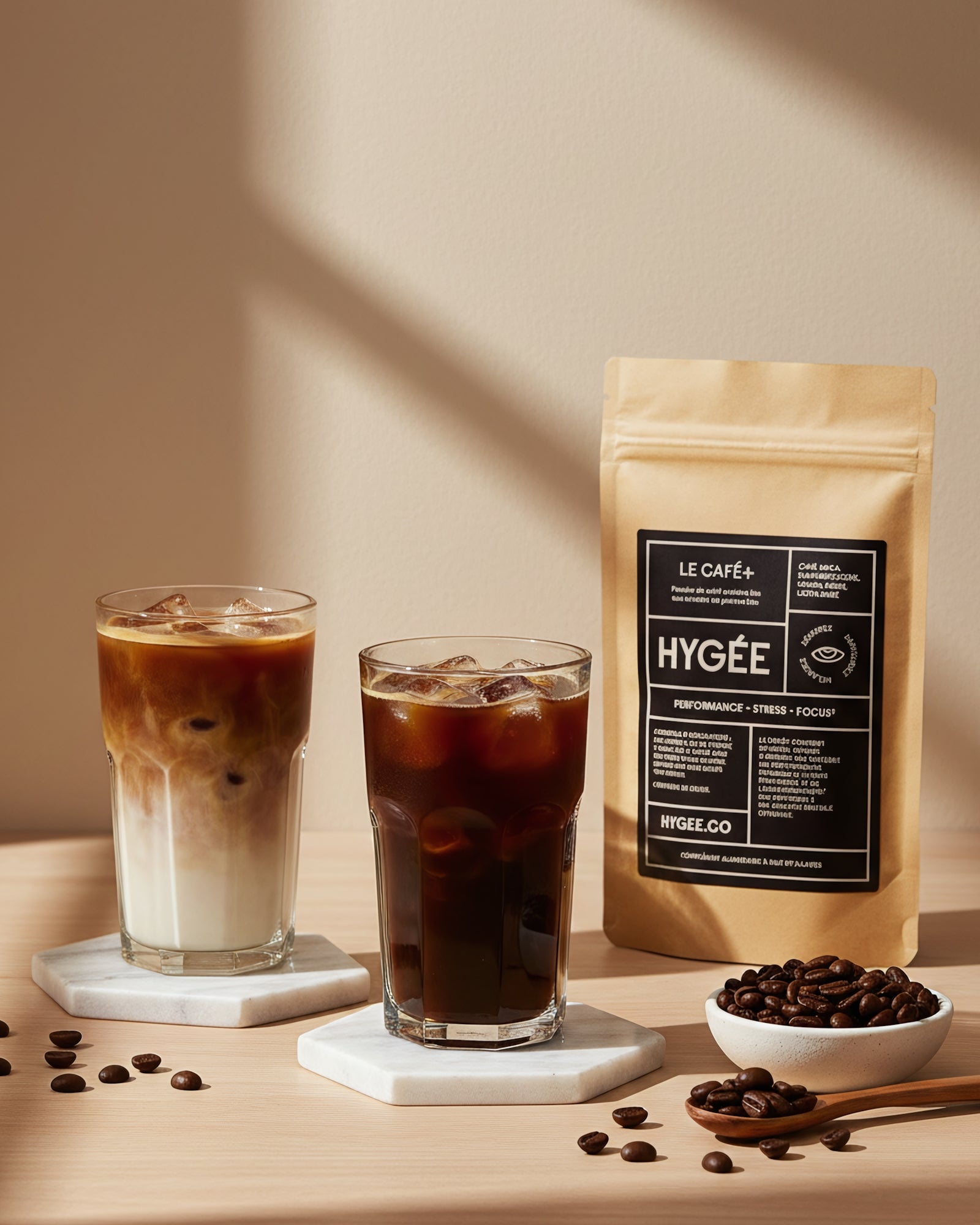 Two tall iced coffees—one layered latte and one black—sit on white hex coasters beside a kraft HYGÉE Le Café+ pouch and a small dish of beans in warm diagonal sunlight. A clean countertop scene with crisp shadows and rich coffee tones.