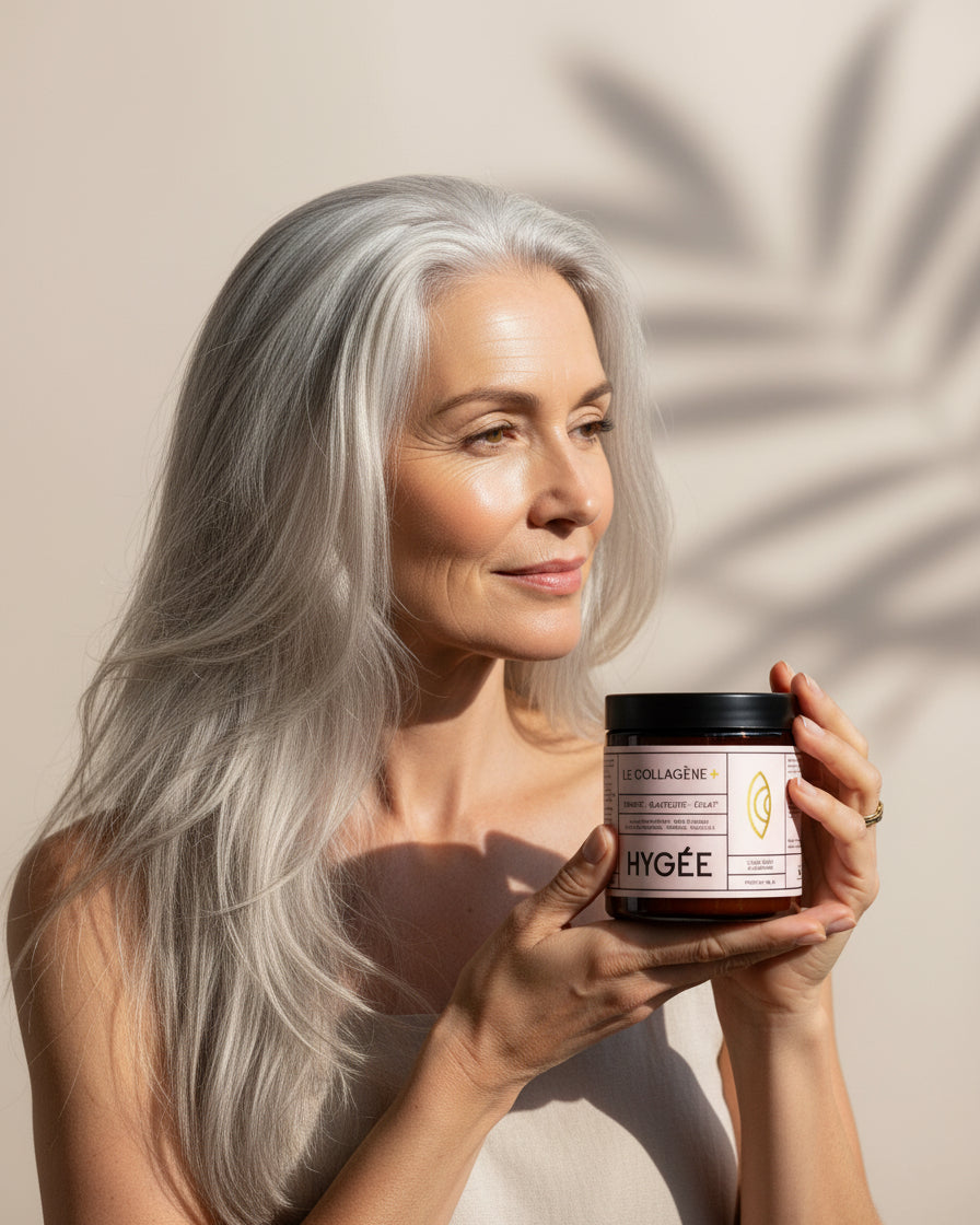 A woman with luminous silver hair presents Allnord’s HYGÉE Le Collagène+ jar in warm daylight, with a soft palm‑leaf shadow on the wall behind. A polished beauty portrait in clean, sun‑washed tones.