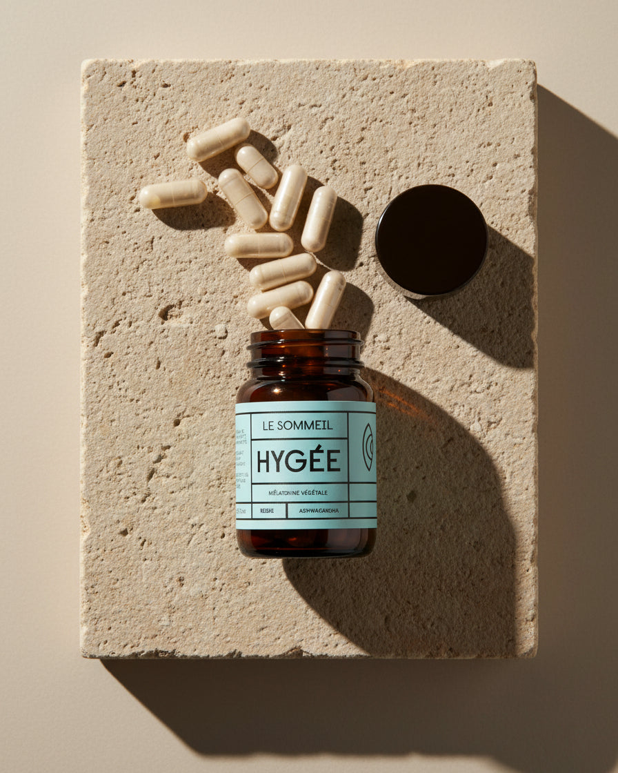 A pastel blue‑label HYGÉE Le Sommeil jar lies open on a warm travertine slab as a scatter of beige capsules spills across the surface with long afternoon shadows.