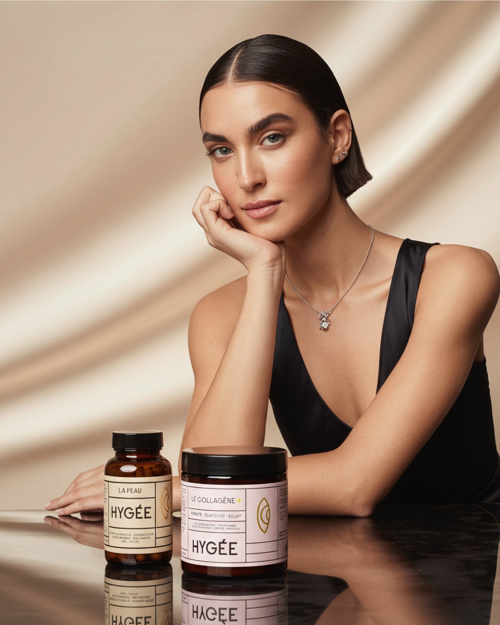 Elegant woman in a black dress sits at a glossy table beside HYGÉE Le Collagène+ collagen powder and La Peau skin capsules, evoking a premium inner‑beauty routine for firmness, radiance and complexion support.