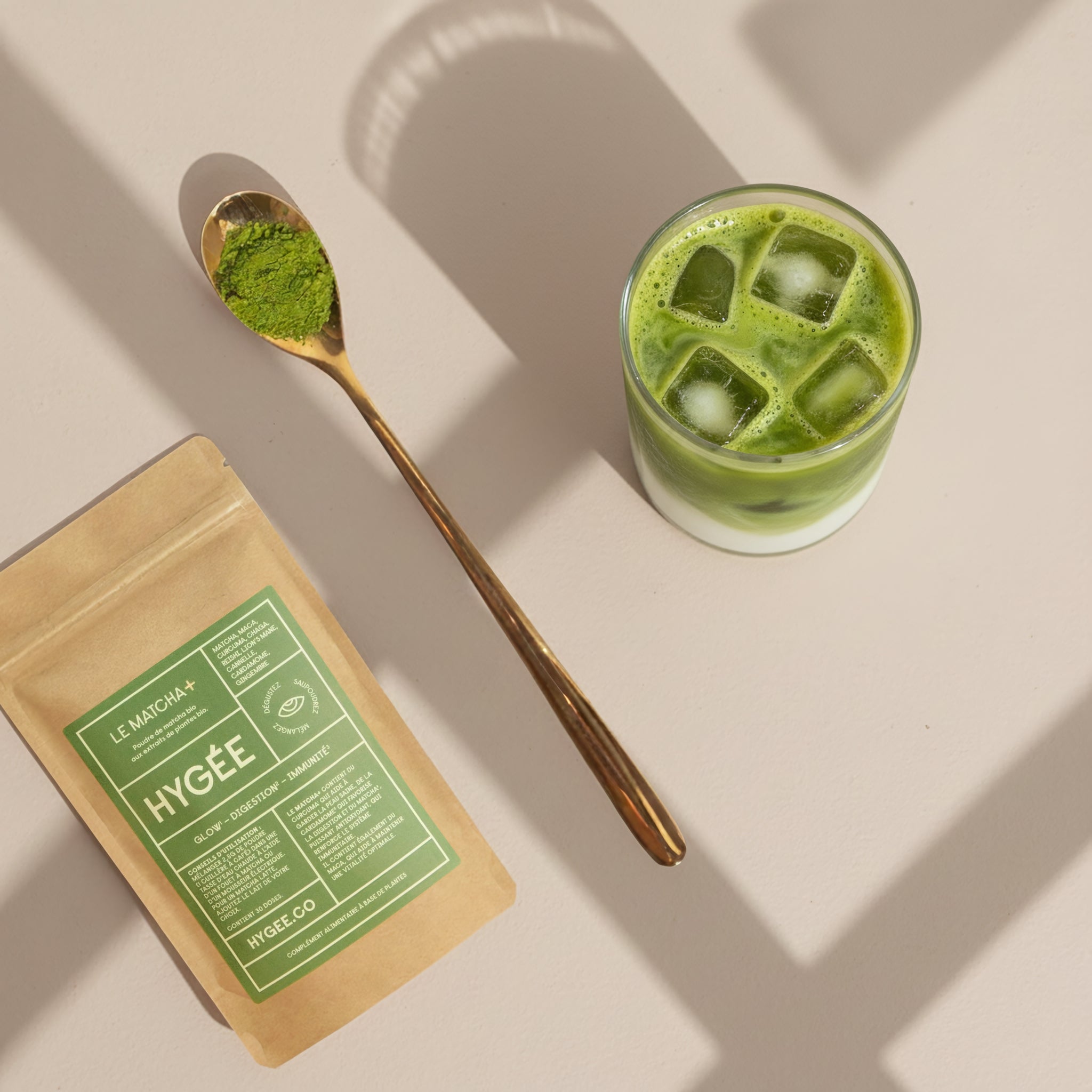 Overhead view of HYGÉE Le Matcha glow and digestion powder pouch beside a golden spoon filled with vibrant green matcha and a layered iced matcha latte on a sunlit neutral surface, highlighting a daily beauty and wellness ritual.