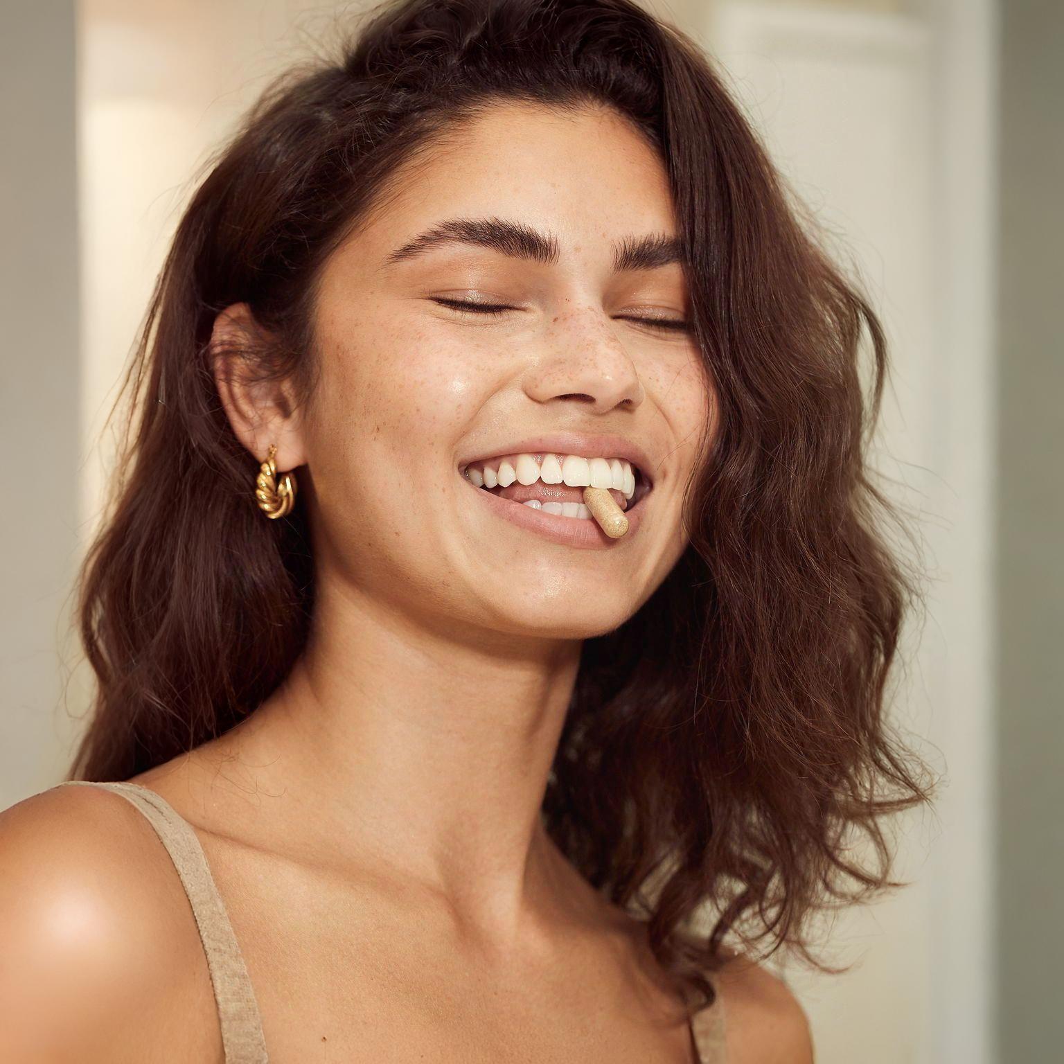 A radiant woman with natural freckles and a genuine smile, holding a Hygée La Chevelure supplement capsule between her teeth, representing a joyful daily wellness ritual.