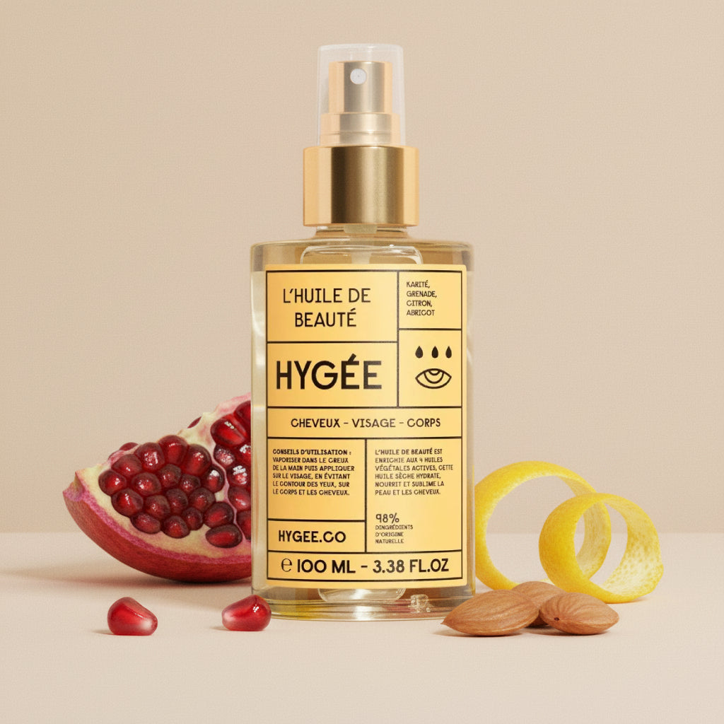 Square glass HYGÉE beauty oil with a gold spray pump stands on a neutral set, styled with a halved pomegranate and scattered arils, lemon peel spirals, and whole almonds in soft daylight.