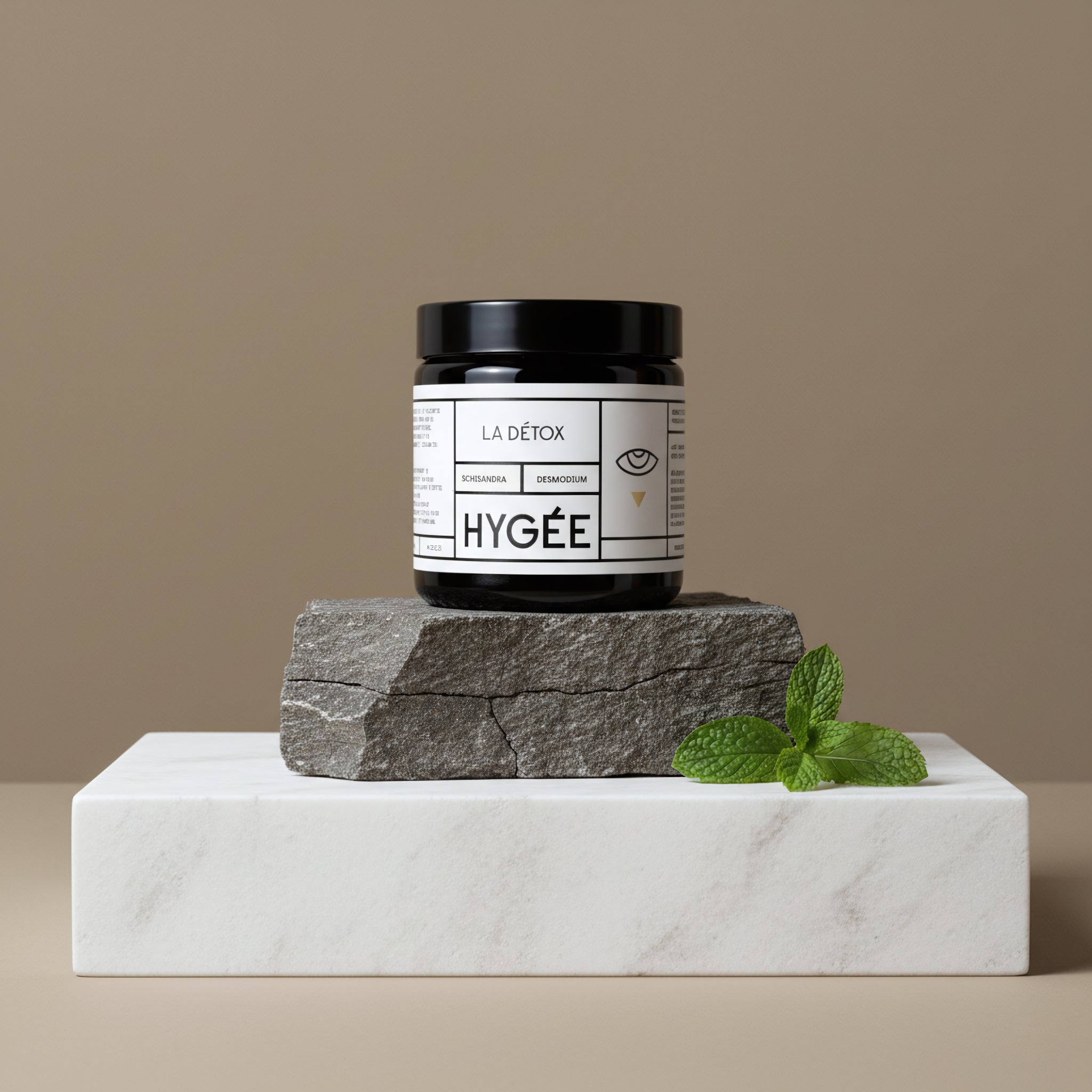 A black‑lidded amber HYGÉE La Détox jar stands centered on a rough gray stone atop a white marble block with a fresh mint sprig, set against a taupe studio backdrop in clean directional light.
