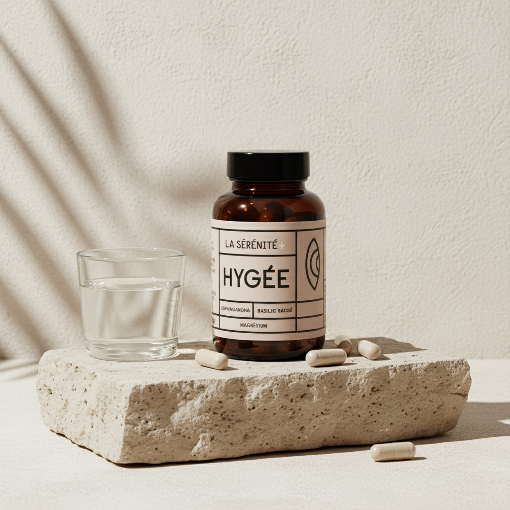 An amber HYGÉE La Sérénité+ bottle stands on a textured stone block beside a clear glass of water, with beige capsules scattered on the surface and soft diagonal shadows on a cream wall.