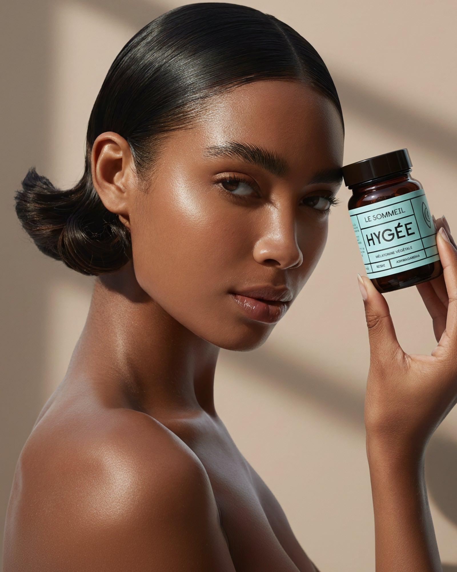 A person with dewy, evening‑light makeup holds an amber HYGÉE Le Sommeil jar with a pastel blue label beside the face against a warm neutral backdrop with soft shadows.