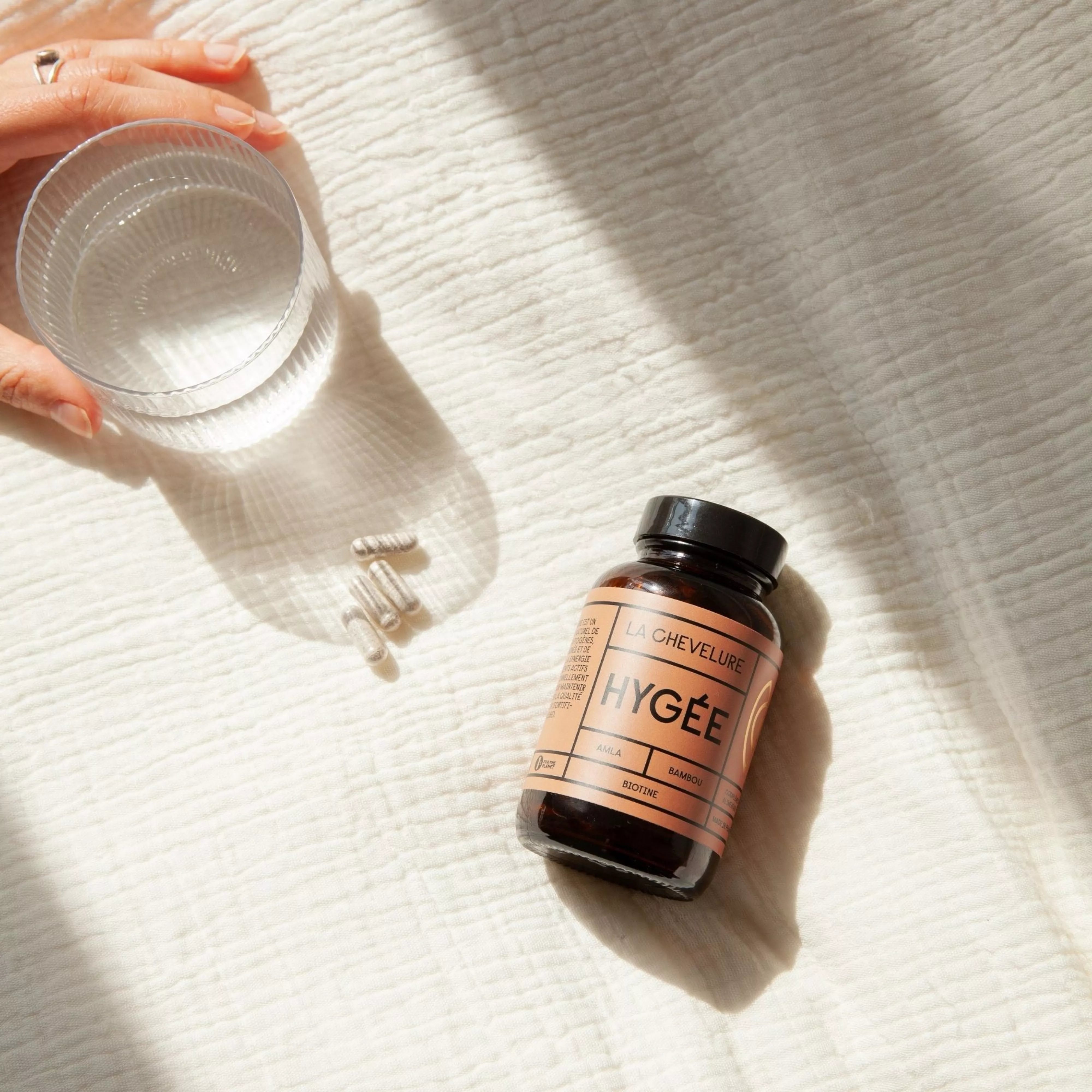 An overhead shot of the Hygée La Chevelure bottle on a textured white cloth, alongside a glass of water and several capsules, illustrating the simple daily discipline of the hair wellness ritual.