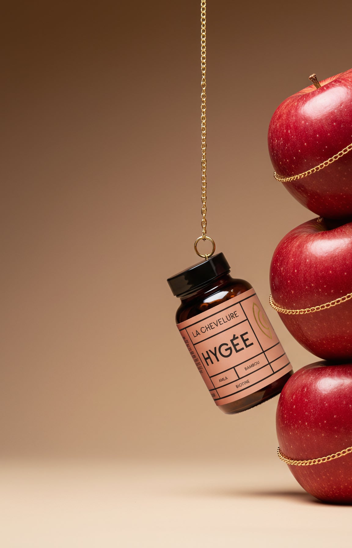 HYGÉE La Chevelure amber supplement bottle for hair strength and growth is suspended from a fine gold chain next to a vertical stack of shiny red apples on a soft beige backdrop, highlighting nourishment, shine, and vitality.