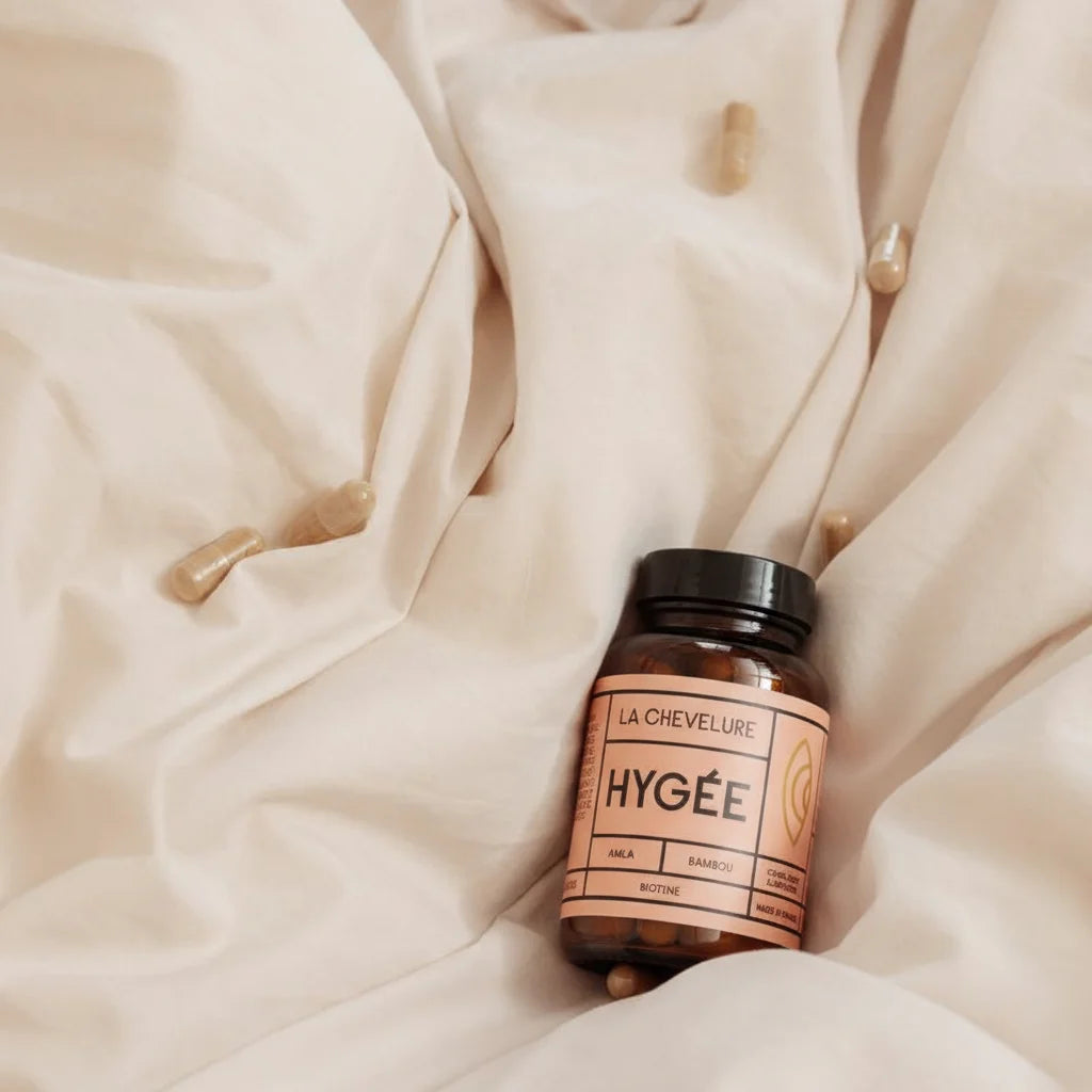 The Hygée La Chevelure bottle nestled in soft, neutral-toned linens with capsules scattered nearby, creating an intimate scene of a morning or evening hair care ritual.
